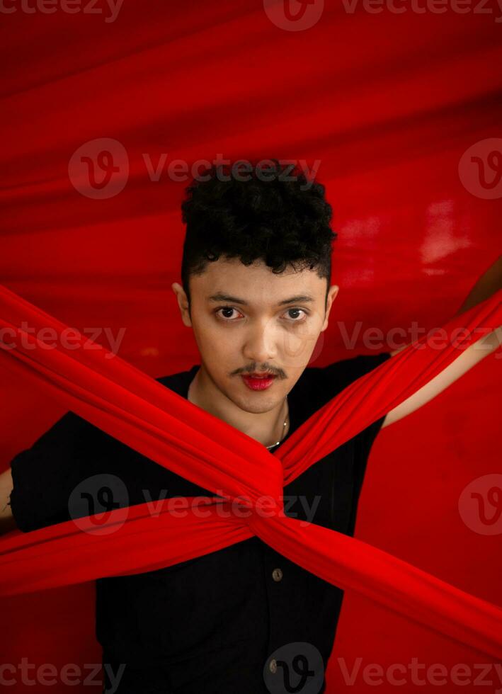 an Asian man stands in front of a red x-shaped cloth and holds it with both hands photo