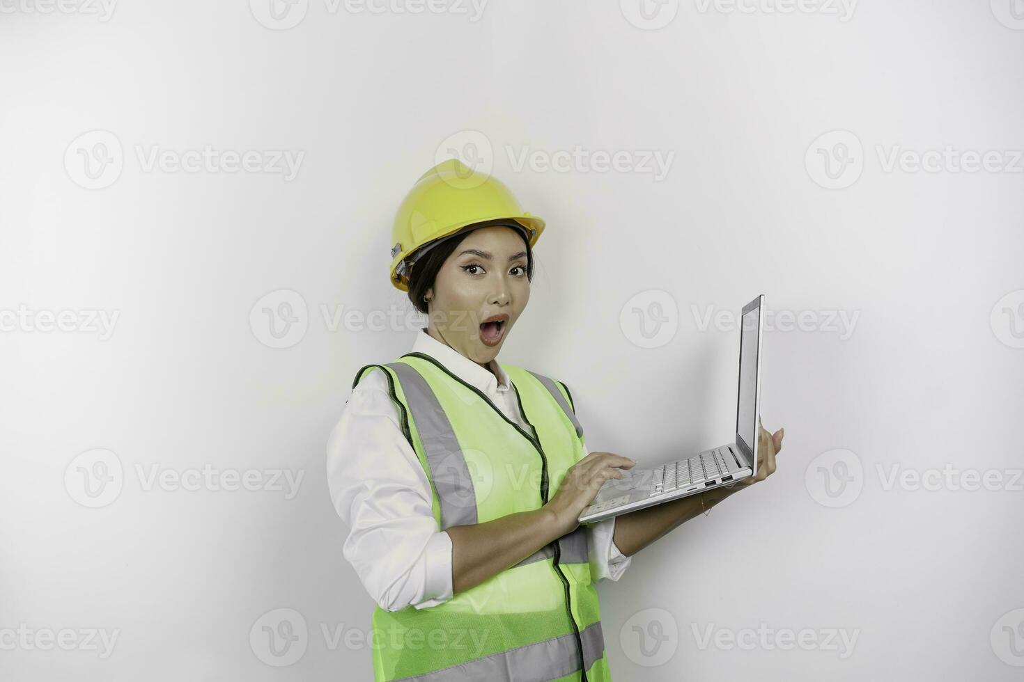 A portrait of a shocked Asian woman labor wearing safety helmet and vest while holding her laptop with her mouth wide open, isolated by white background. Labor's day concept photo
