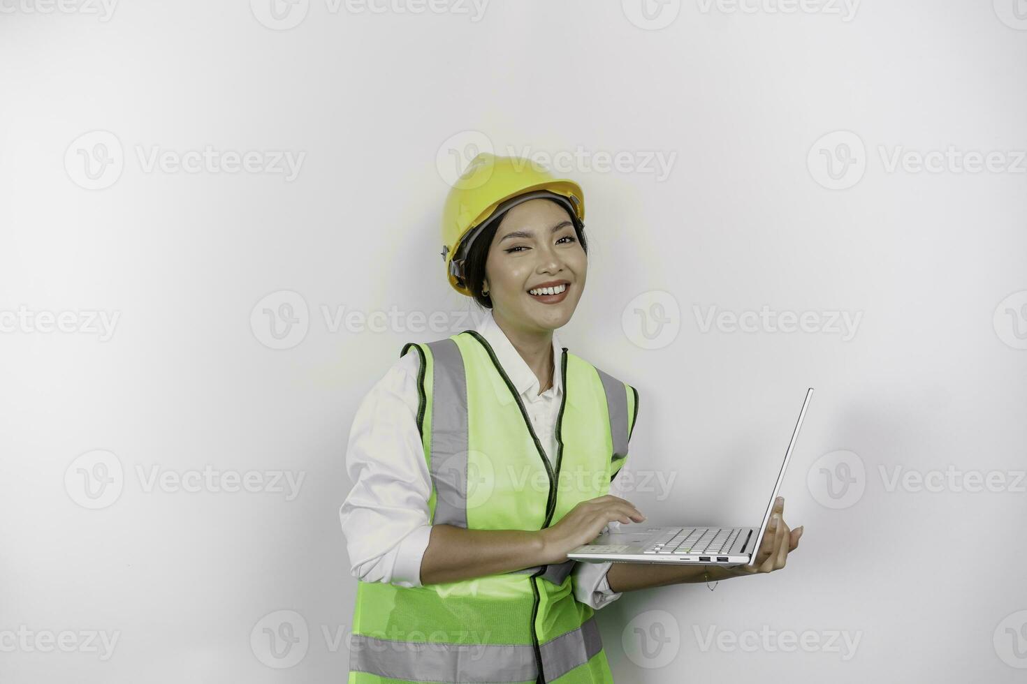 A smiling Asian woman labor wearing safety helmet and vest while holding her laptop, isolated by white background. Labor's day concept. photo