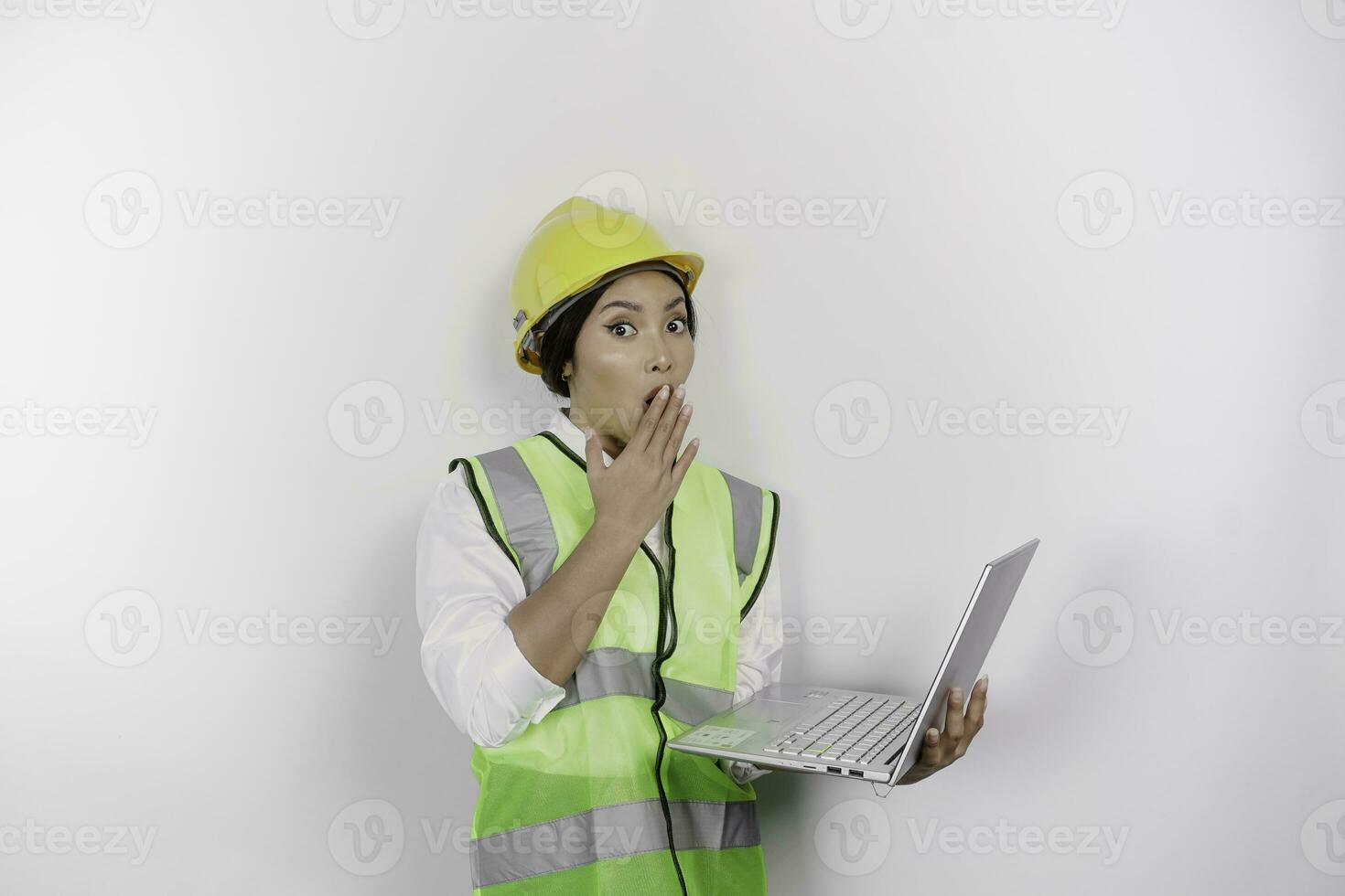 A portrait of a shocked Asian woman labor wearing safety helmet and vest while holding her laptop with her mouth wide open, isolated by white background. Labor's day concept photo