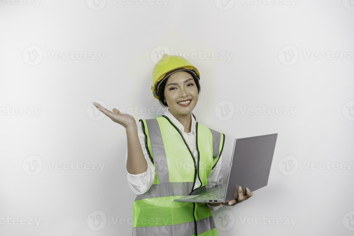 A smiling Asian woman labor wearing safety helmet and vest, pointing to copy space above her and holding her laptop, isolated by white background. Labor's day concept. photo