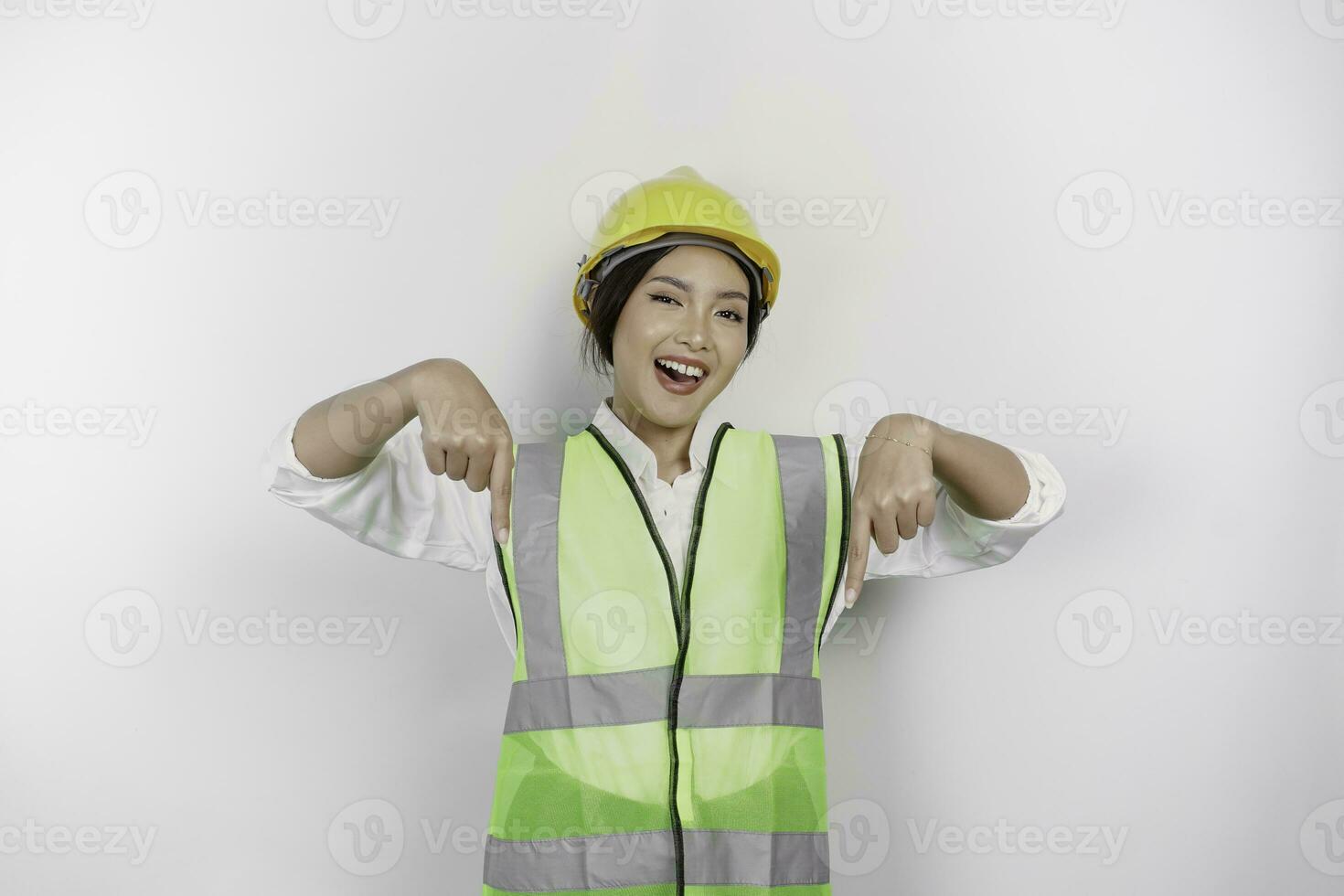 A smiling Asian woman labor wearing safety helmet and vest, pointing to copy space below her, isolated by white background. Labor's day concept. photo