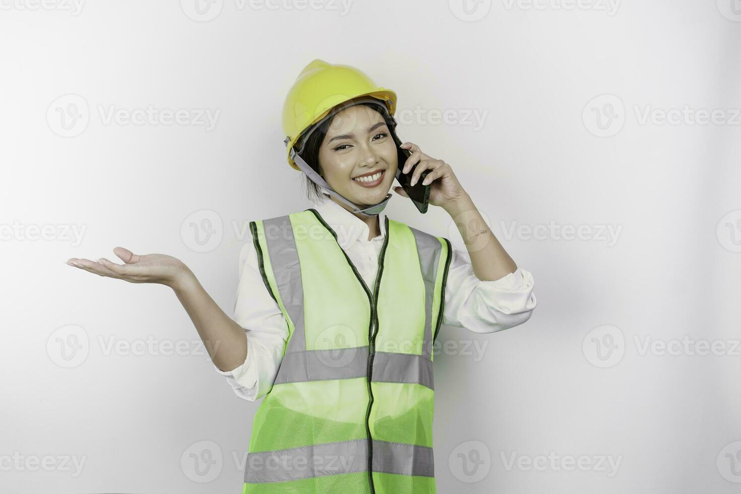A smiling Asian woman labor wearing safety helmet and vest, having a phone call and pointing to copy space beside her, isolated by white background. Labor's day concept. photo