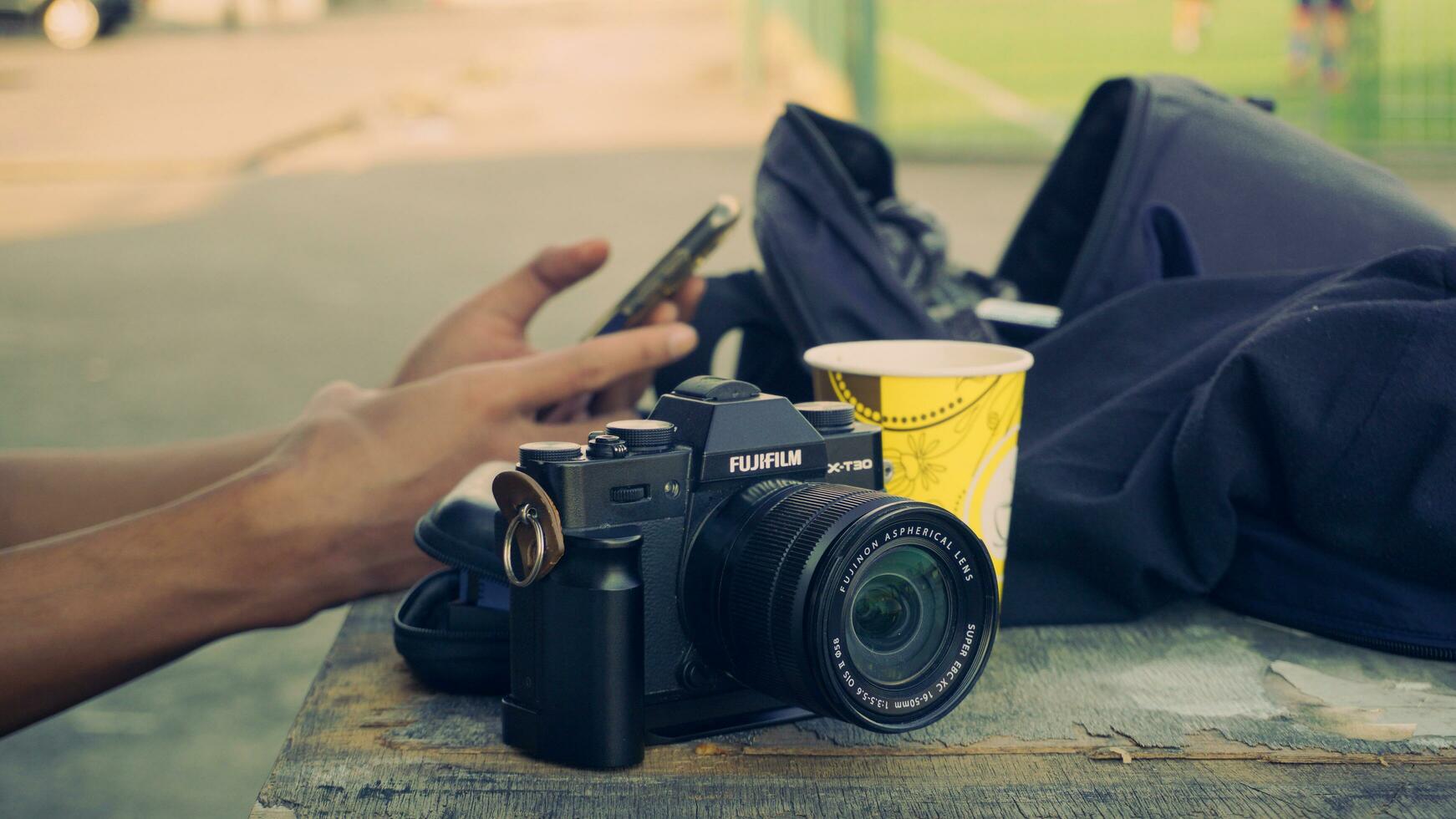 Fujifilm X T30 camera placed on a wooden surface with a bag in the background and a hand holding a smartphone. photo