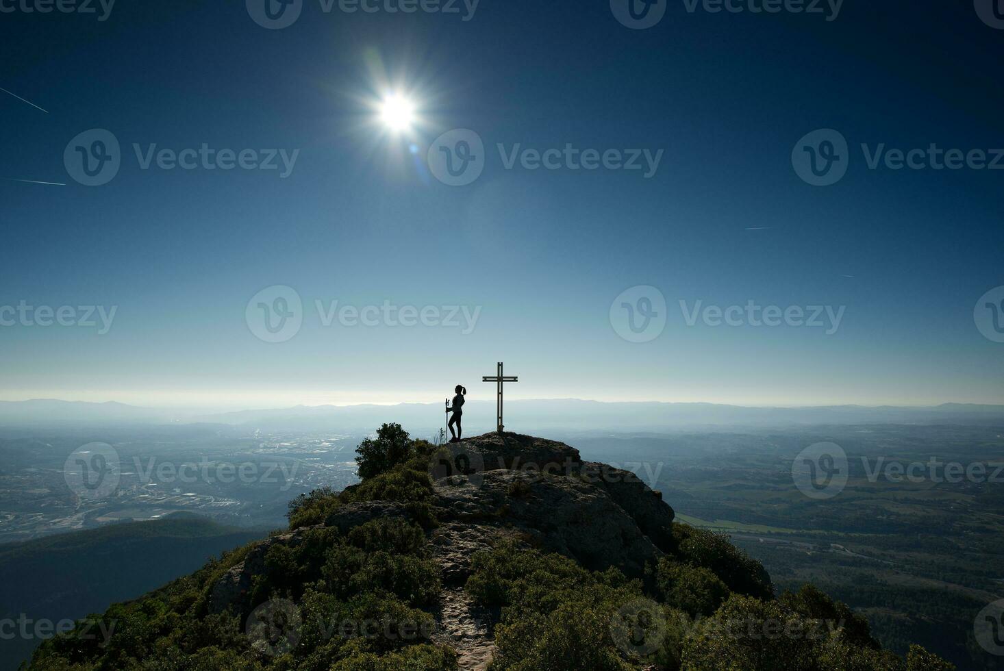 Woman contemplating a backlit landscape from the top of a hill, where there is a cross. photo