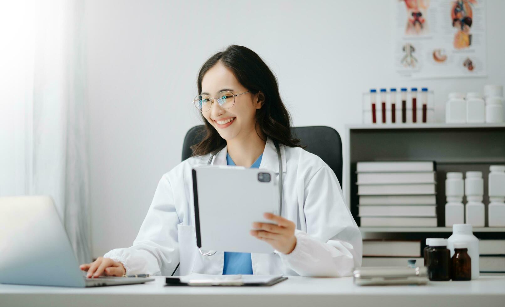 Beautiful physician doctor sit on table in office hospital. Attractive therapist female practitioner pharmacist smile and use tablet and laptop. after success work from treatment photo