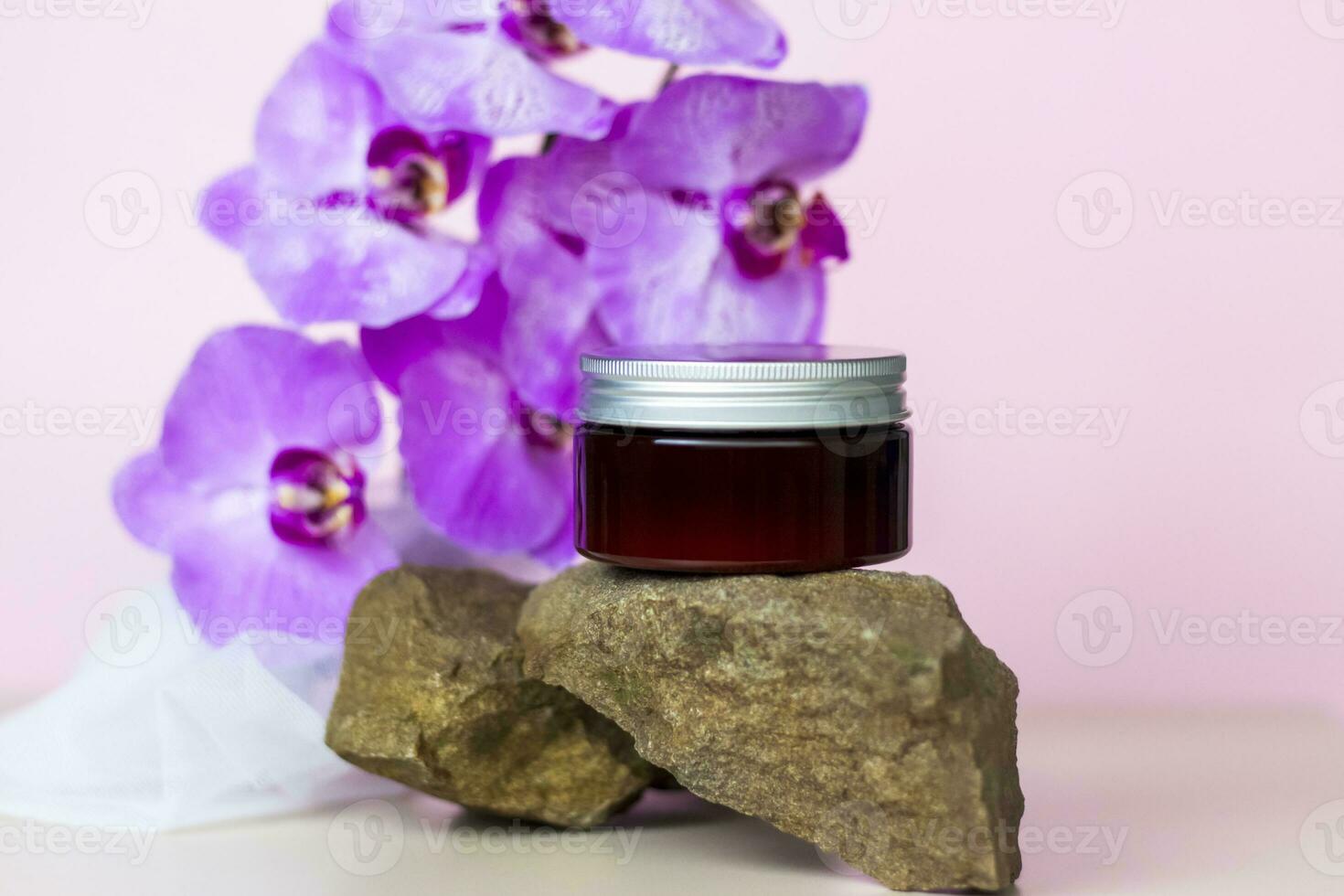 A brown jar of cream on a pink background stands on a large building stone with beautiful orchids next to it. Stylish appearance of the product, layout, personality. photo