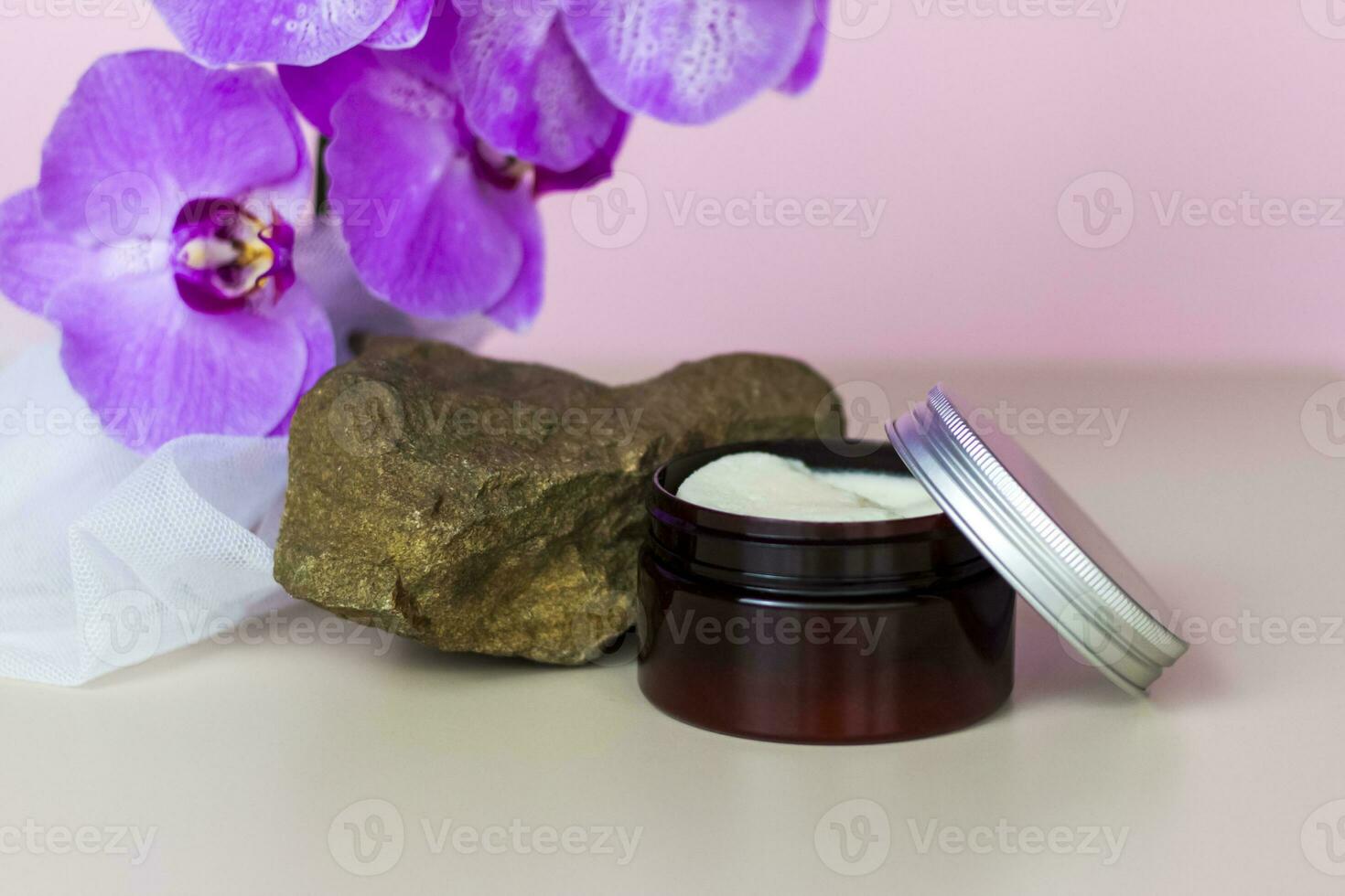 A brown jar of cream on a pink background stands on a large building stone with beautiful orchids next to it. Stylish appearance of the product, layout, personality. photo