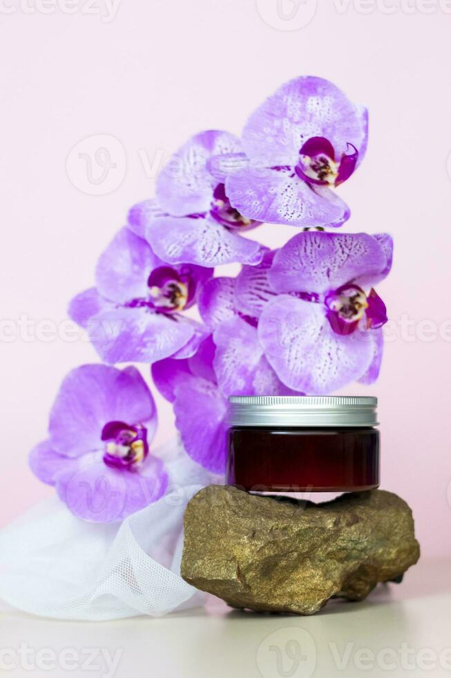 A brown jar of cream on a pink background stands on a large building stone with beautiful orchids next to it. Stylish appearance of the product, layout, personality. photo