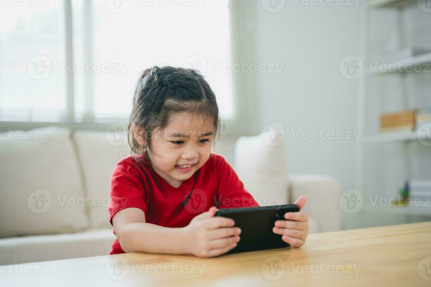 Asian child girl looking using and touch mobile phone screen. Baby smiling funny time to use mobile phone. Too much screen time. Cute girl watching videos while tv in living room, Internet addiction. photo