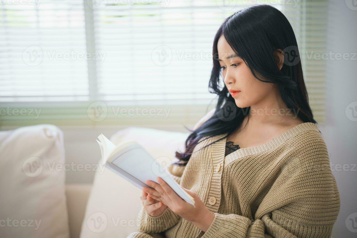 Joyful happy asian woman smiling and reading book while sitting on couch sofa in living room at home. Asian women relax reading book smile at sofa In the house. Activity hobby at house concept. photo