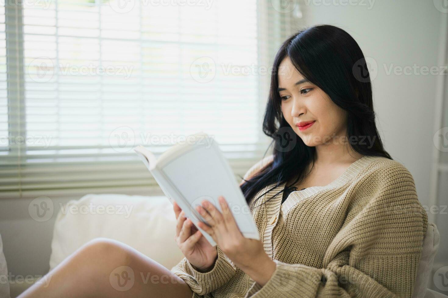 Joyful happy asian woman smiling and reading book while sitting on couch sofa in living room at home. Asian women relax reading book smile at sofa In the house. Activity hobby at house concept. photo