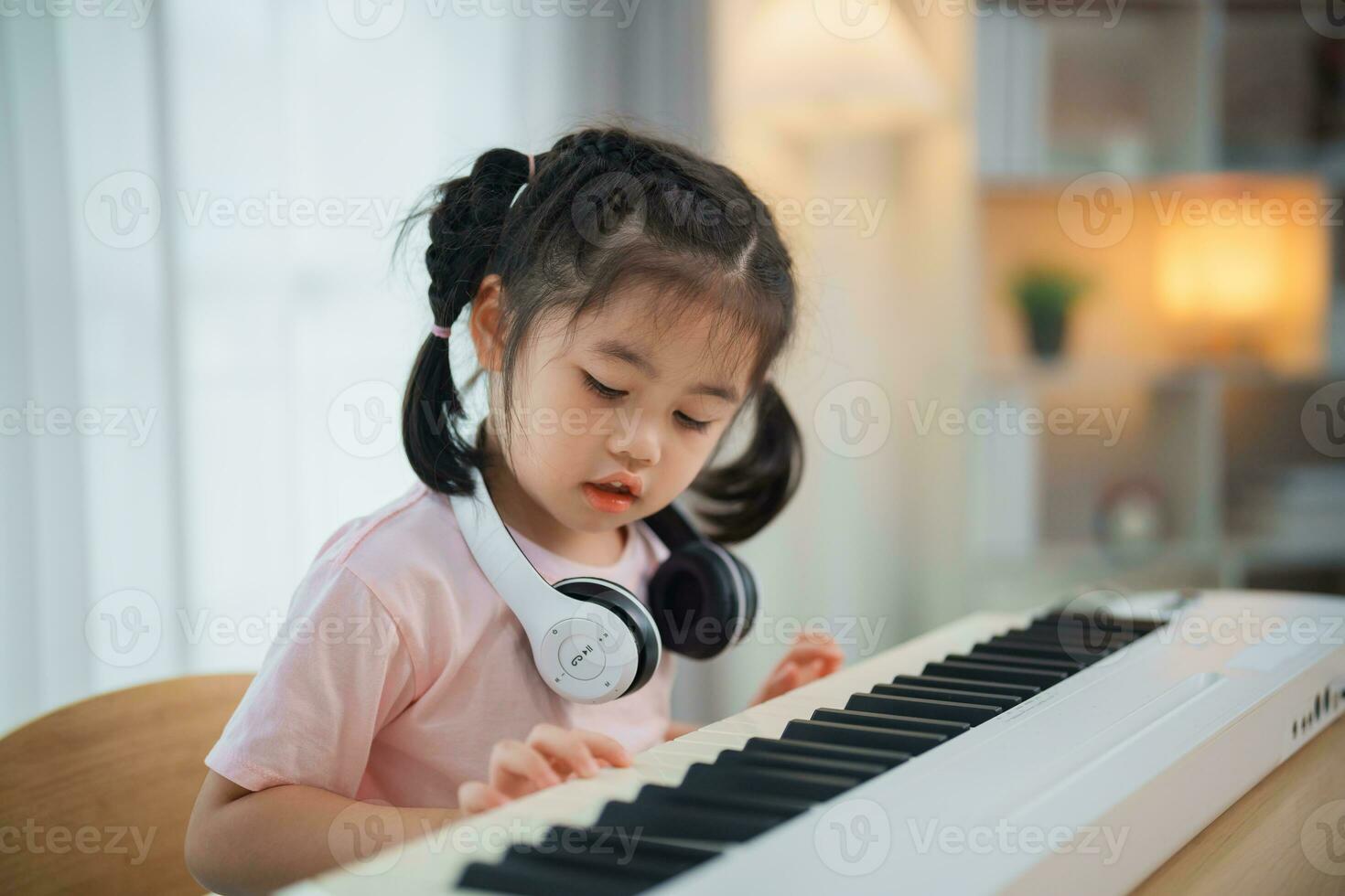 Asian cute girl smile and wearing white headphone playing learning online piano music in the living room at home. The idea of activities for the child at home during quarantine. Music learning study. photo