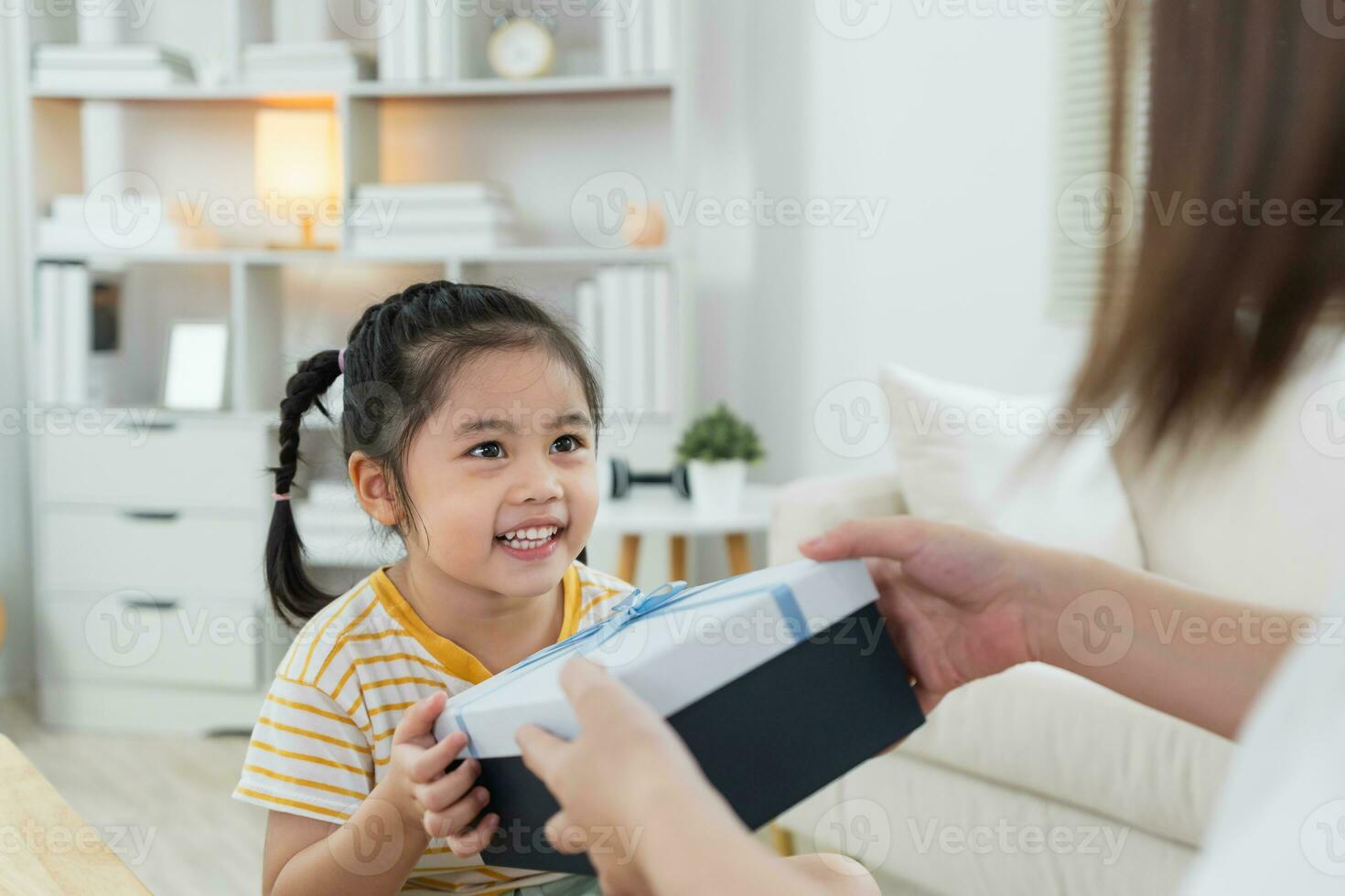 Grateful asian girl holding blue present box giving mothers day gift to mom. Happy mom and cute daughter kid child celebrating birthday, hugging on couch sofa at home. Happy family concept. photo