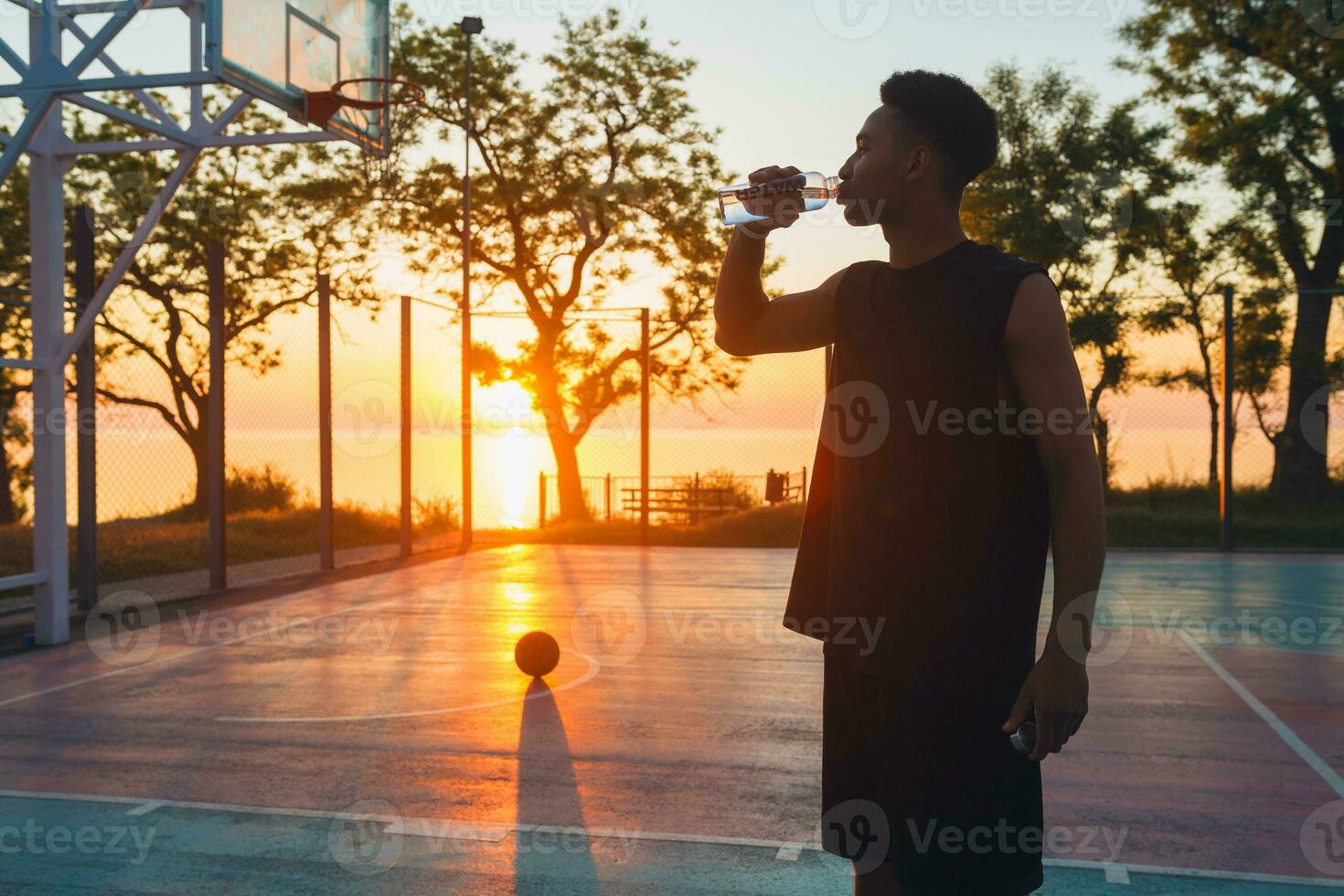 negro hombre haciendo Deportes en mañana, Bebiendo agua en baloncesto Corte en amanecer foto