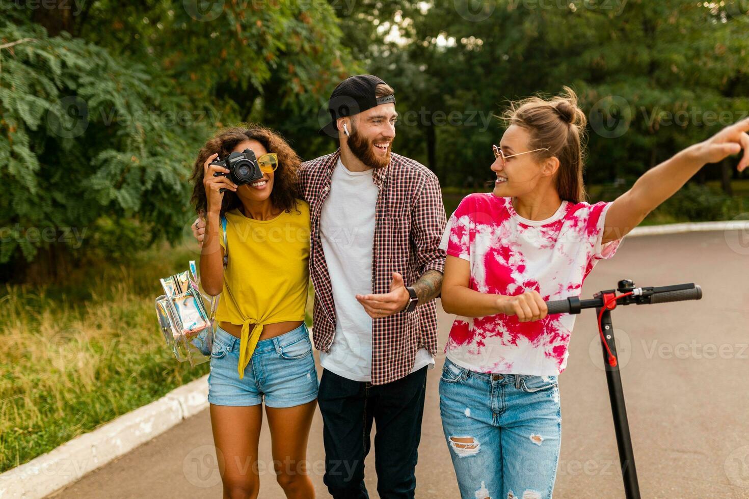 happy young company of smiling friends walking in park with electric kick scooter photo
