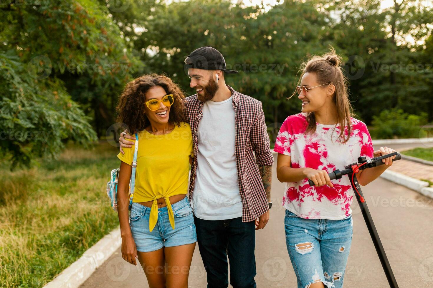 happy young company of smiling friends walking in park with electric kick scooter photo