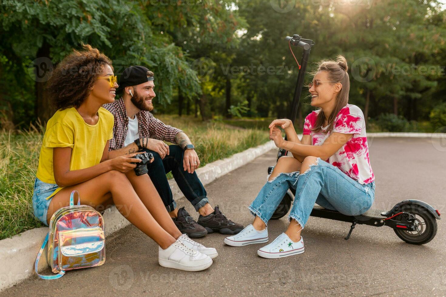 contento joven empresa de sonriente amigos sentado en parque en césped con eléctrico patada scooter foto