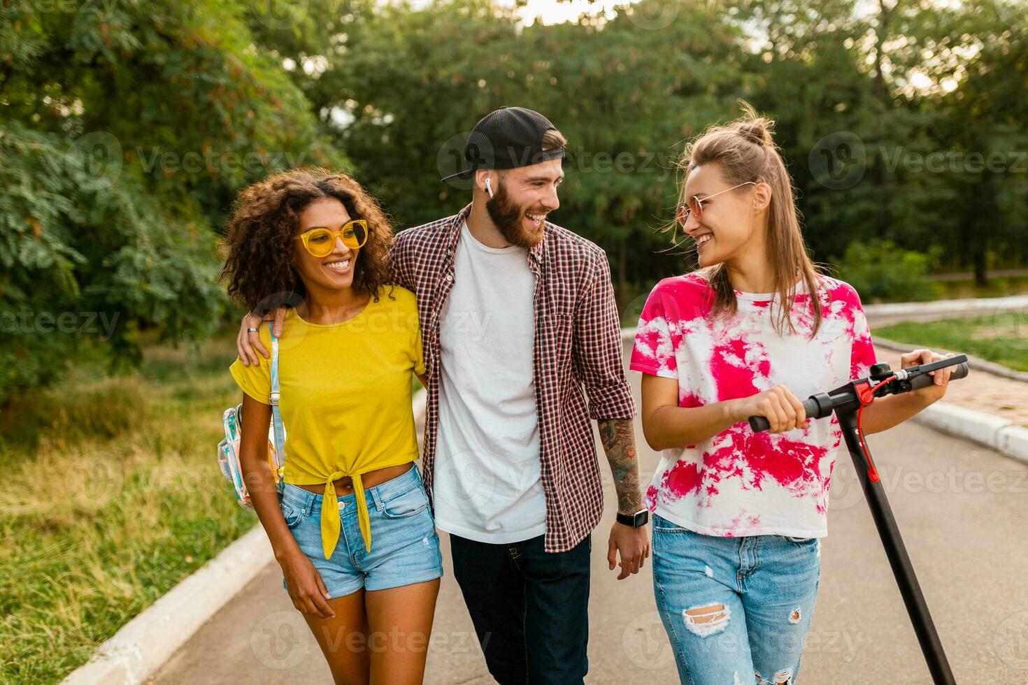 happy young company of smiling friends walking in park with electric kick scooter photo