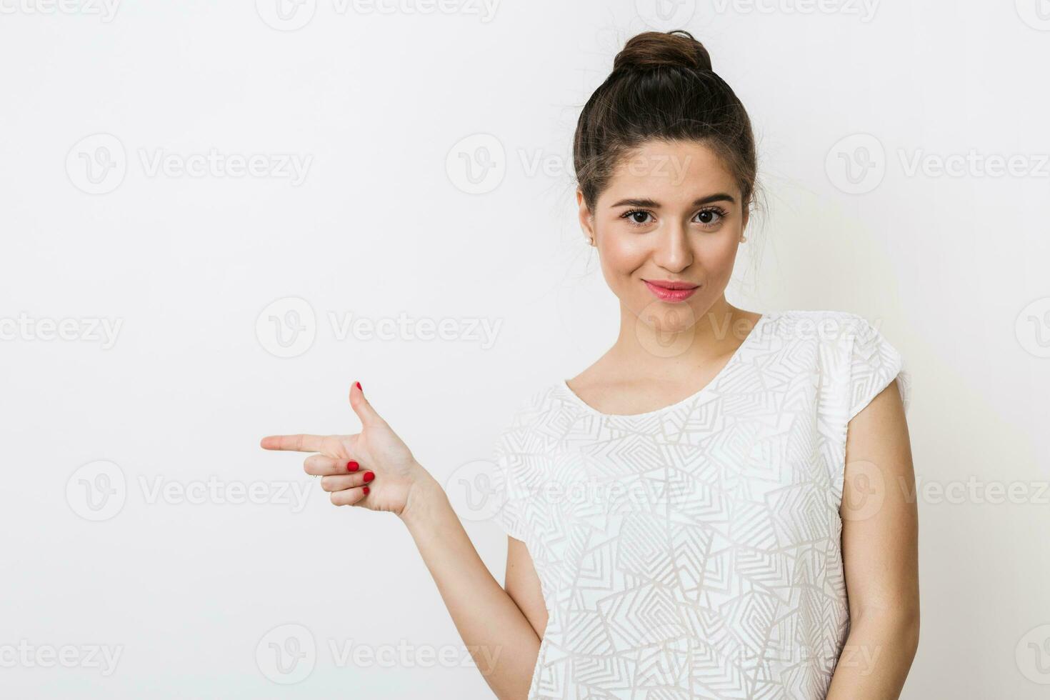 young woman in white blouse pointing finger to the aside, looking into camera, isolated on studio background, tricky smile photo