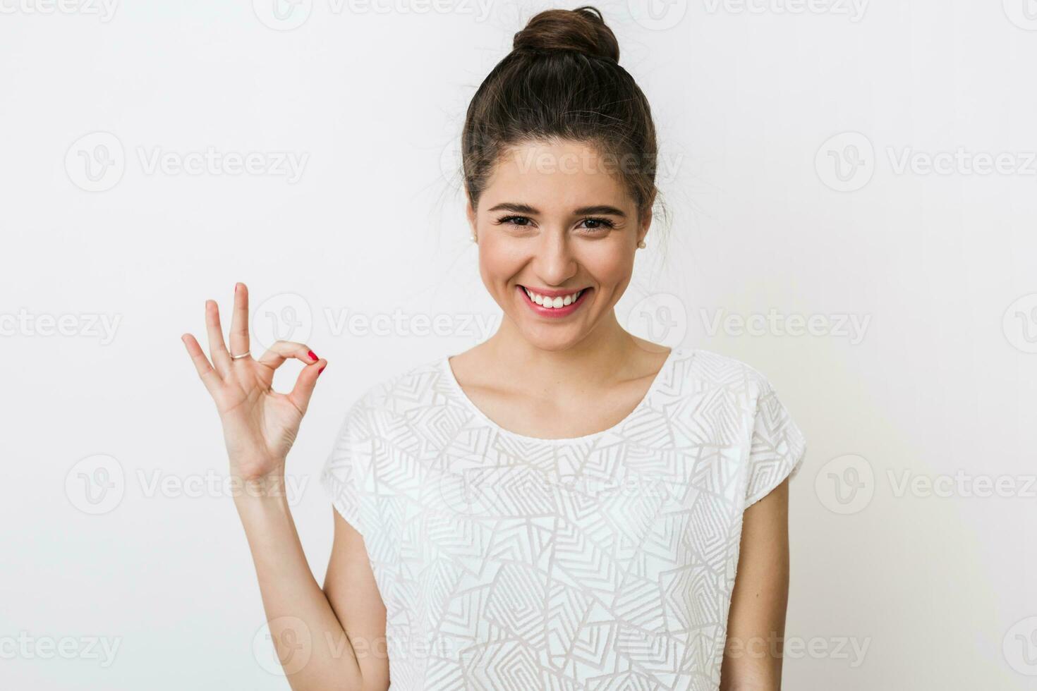 young happy woman in white blouse showing okay sign, positive gesture, looking into camera, smiling, isolated on studio background photo