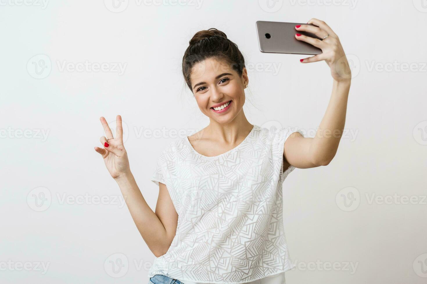 young attractive smiling woman in white blouse holding smart phone, showing peace sign, positive emotion, making selfie photo, isolated on studio background photo