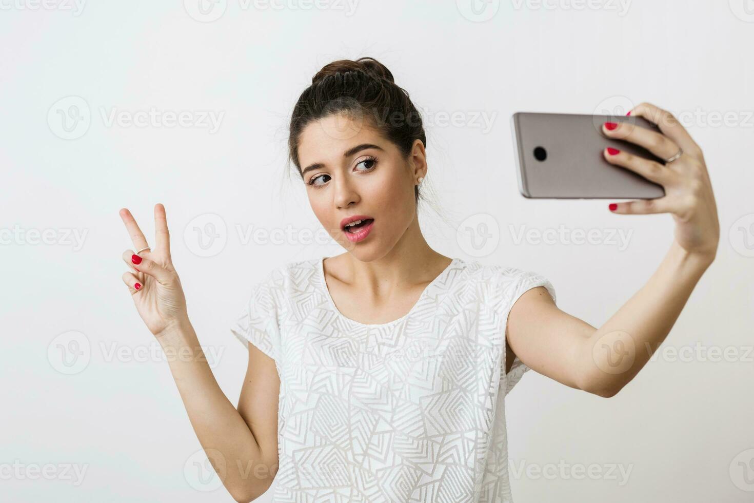 young attractive smiling woman in white blouse holding smart phone, showing peace sign, positive emotion, making selfie photo, isolated on studio background photo