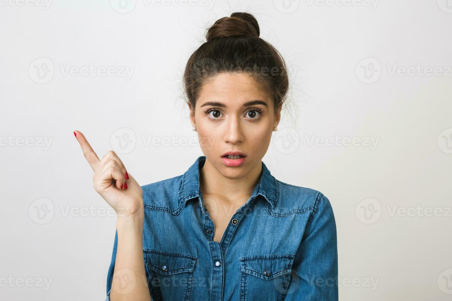 close-up portrait of stylish attractive woman in denim blue shirt with shocked expression of face, pointing finger up, wide opened eyes and mouth, emotional, isolated on white background photo