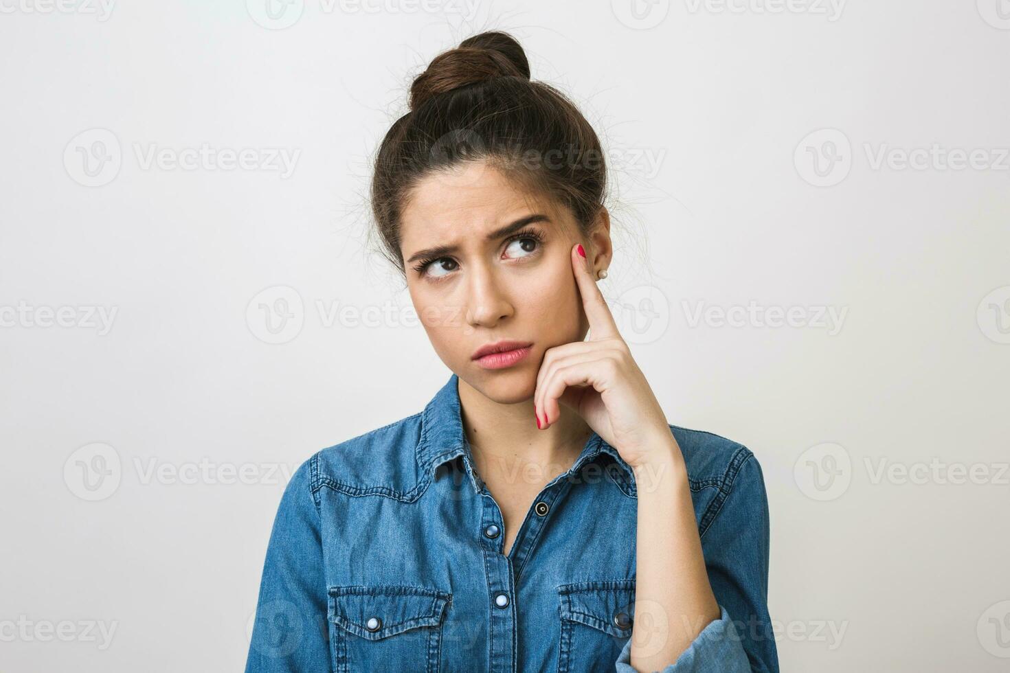 young woman thinking, holding finger at her temple, looking up, stylish denim shirt, isolated on white studio background photo