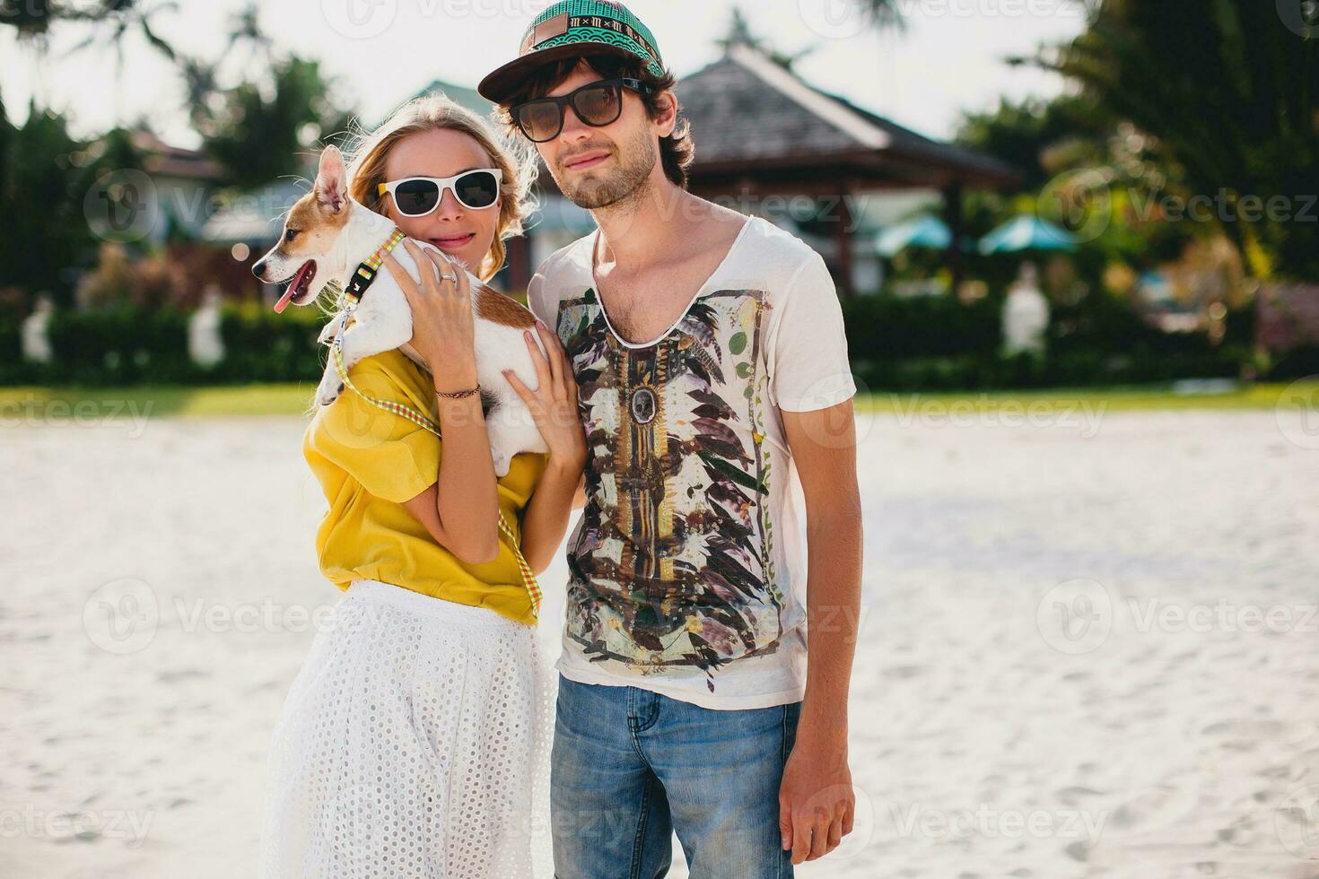 hipster young stylish hipster couple in love walking playing dog puppy jack russell in tropical beach, white sand, cool outfit, romantic mood, having fun, sunny, man woman together, vacation photo
