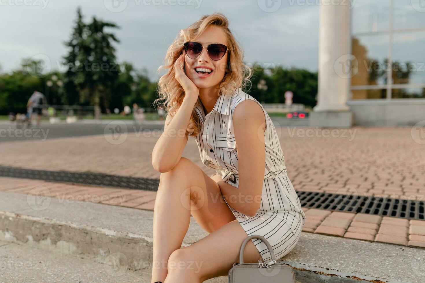 young attractive cute stylish blonde woman sitting in city street in summer fashion style photo