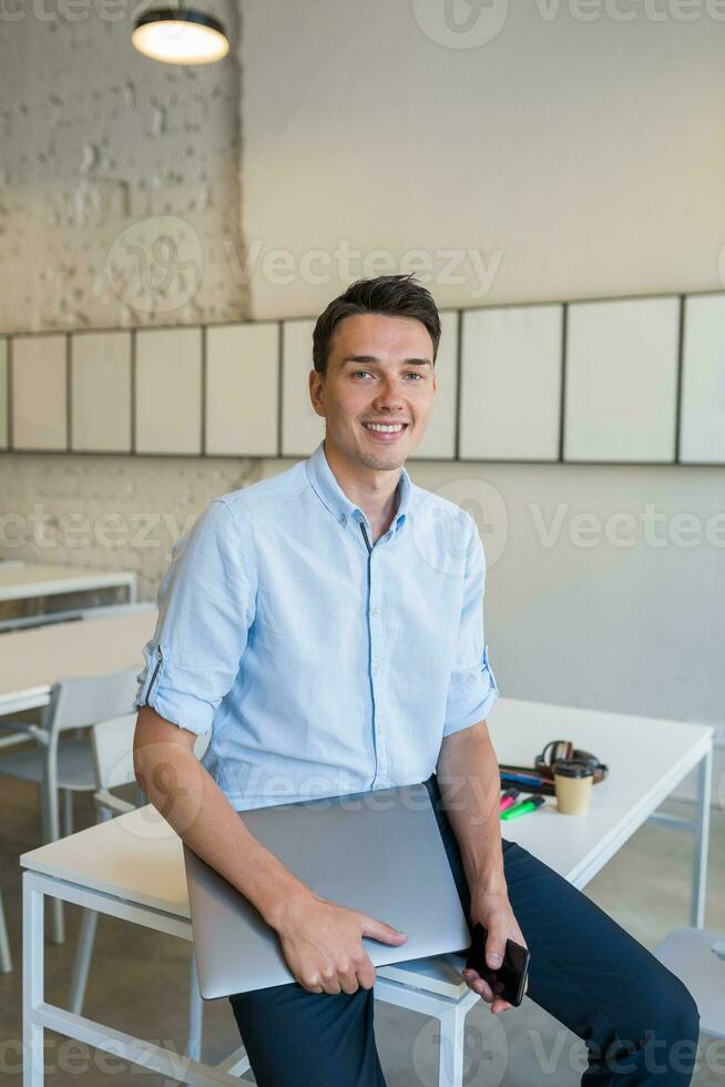 young attractive smiling man sitting in co-working open office, holding laptop photo