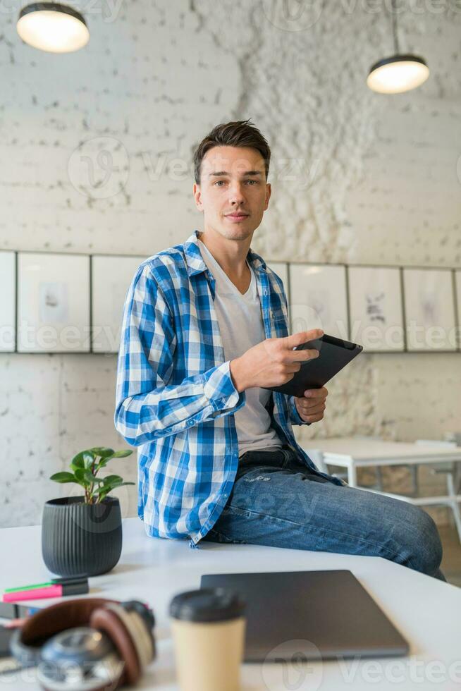 young handsome man in chekered shirt sitting on table using tablet computer in co-working office photo