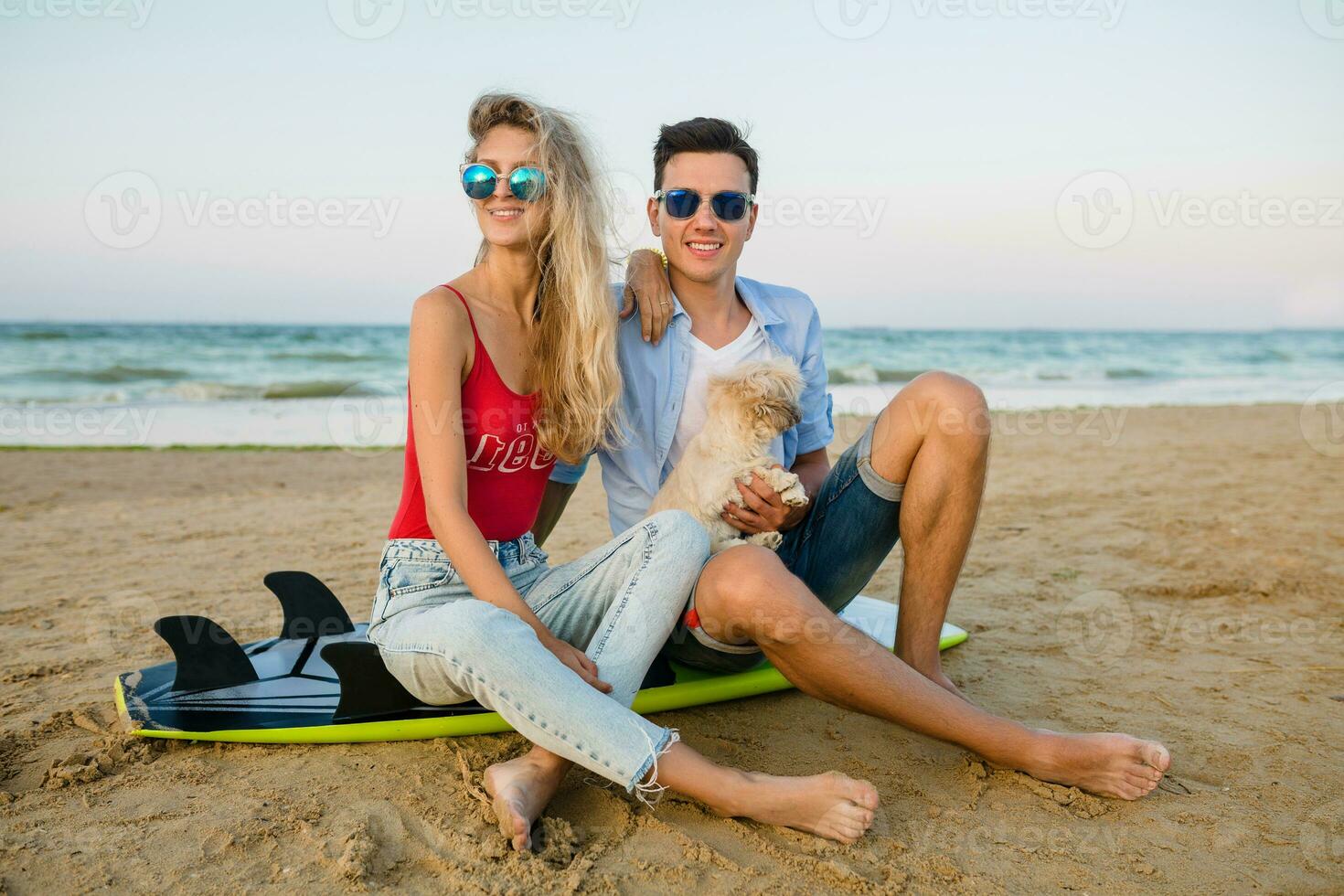 young smiling couple having fun on beach sitting on sand with surf boards playing with dog photo