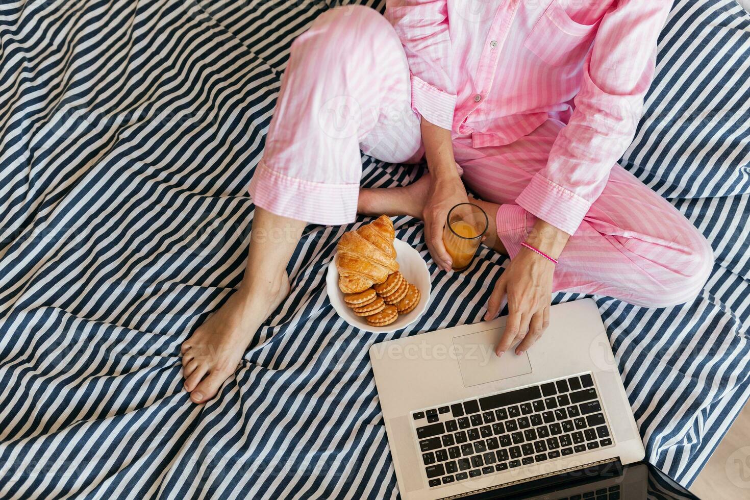cute young pretty blond woman in pink pajamas sitting on bed working on laptop, freelancer at home photo