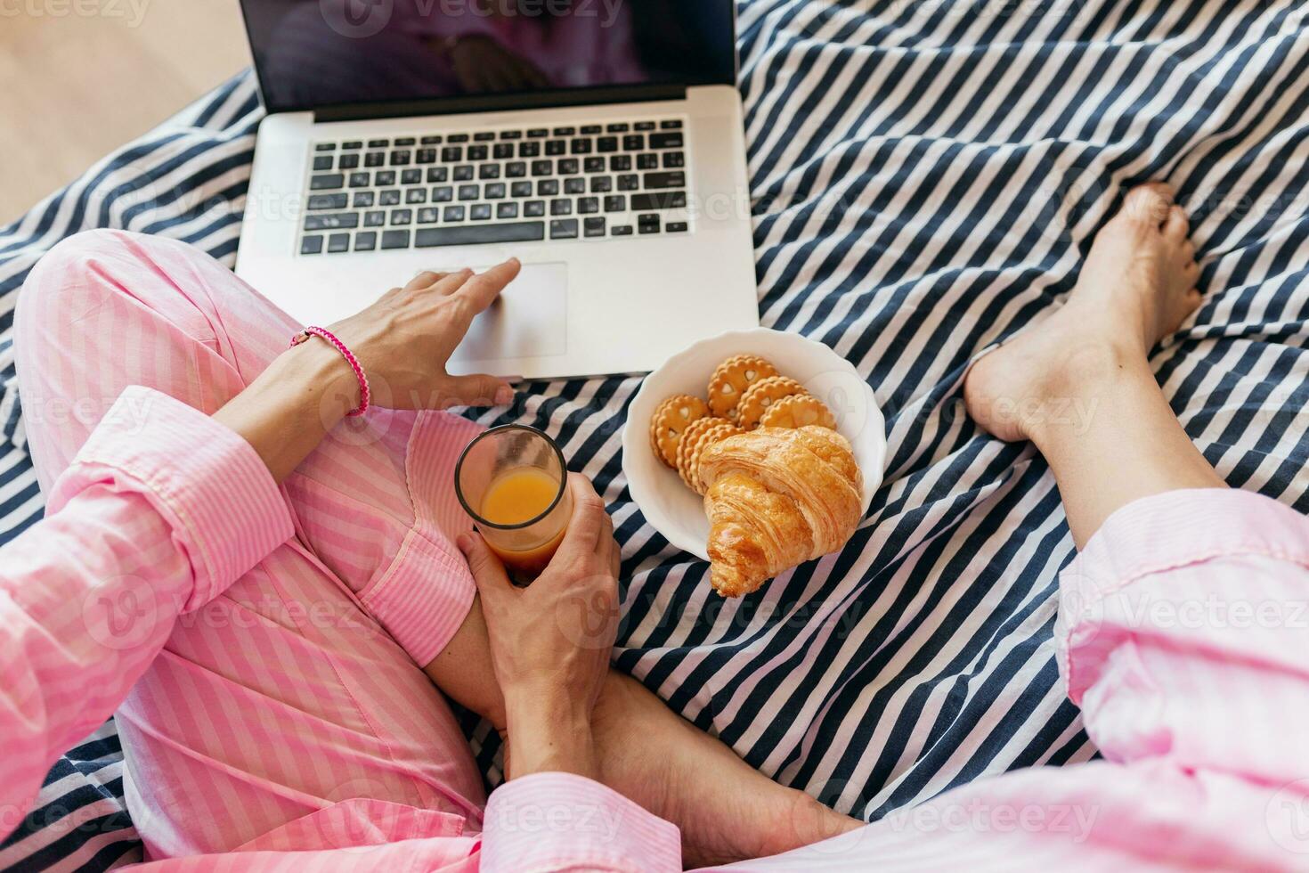 cute young pretty blond woman in pink pajamas sitting on bed working on laptop, freelancer at home photo