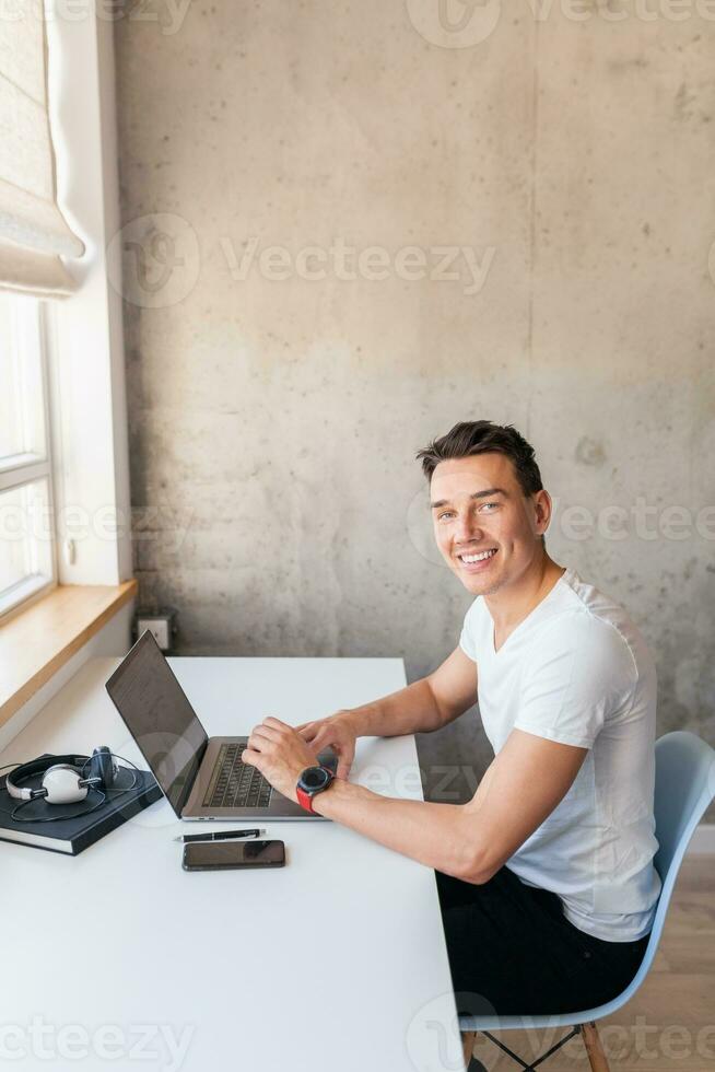 young handsome man in casual outfit sitting at table working on laptop, typing, freelancer at home photo