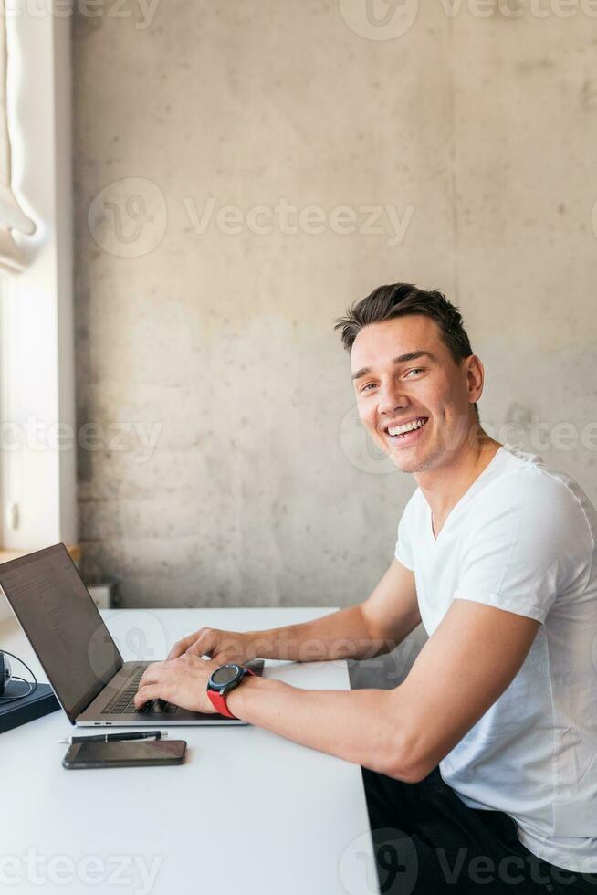 young handsome smiling man in casual outfit sitting at table working on laptop photo