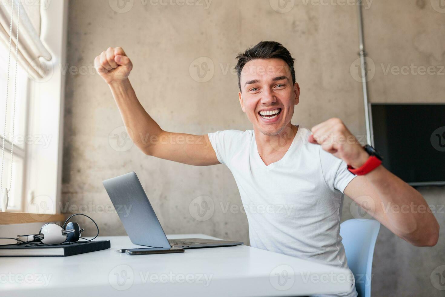 young handsome smiling man in casual outfit sitting at table working on laptop photo