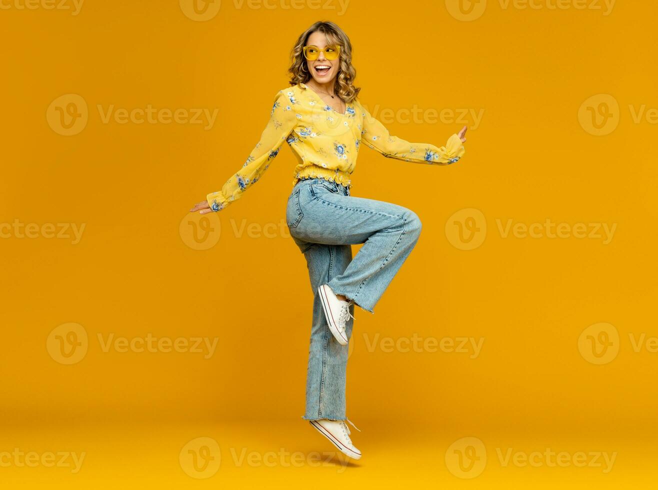 stylish happy smiling blond woman posing in jeans on yellow background photo