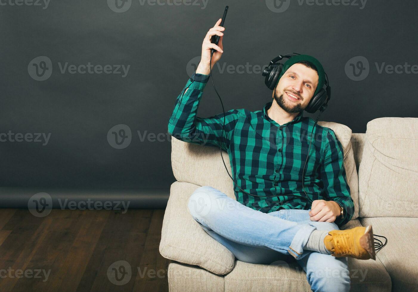 young hipster handsome bearded man sitting on a couch at home, listening to music on headphones photo