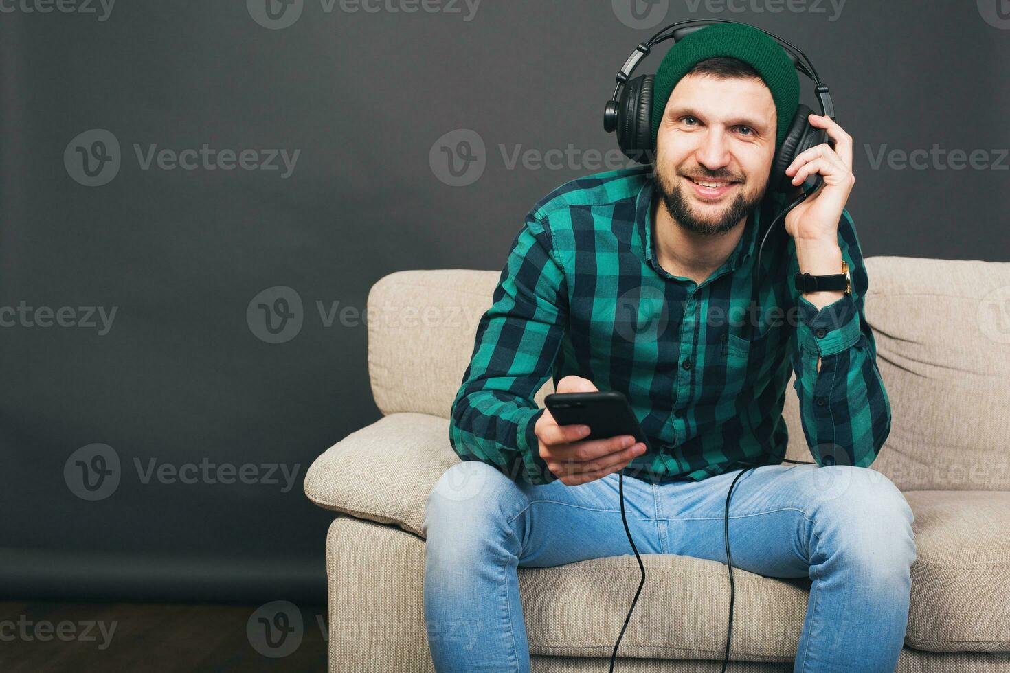 young hipster handsome bearded man sitting on a couch at home, listening to music on headphones photo