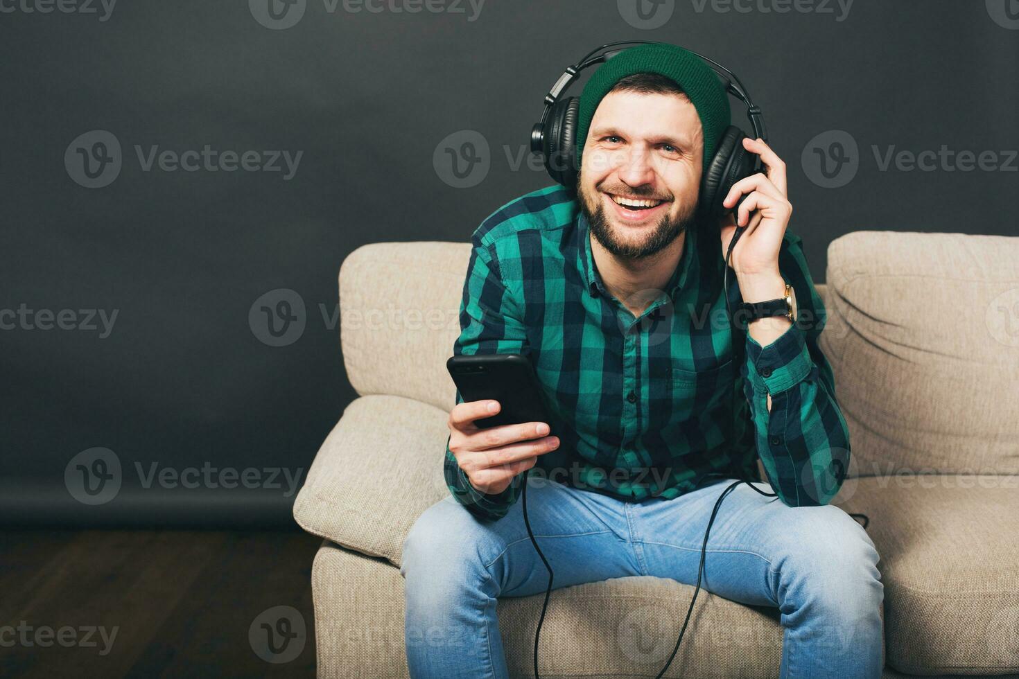 young hipster handsome bearded man sitting on a couch at home, listening to music on headphones photo