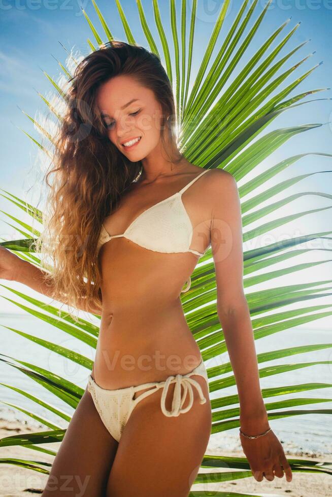 joven flaco mujer en blanco bikini trajes de baño participación hoja de palma árbol foto