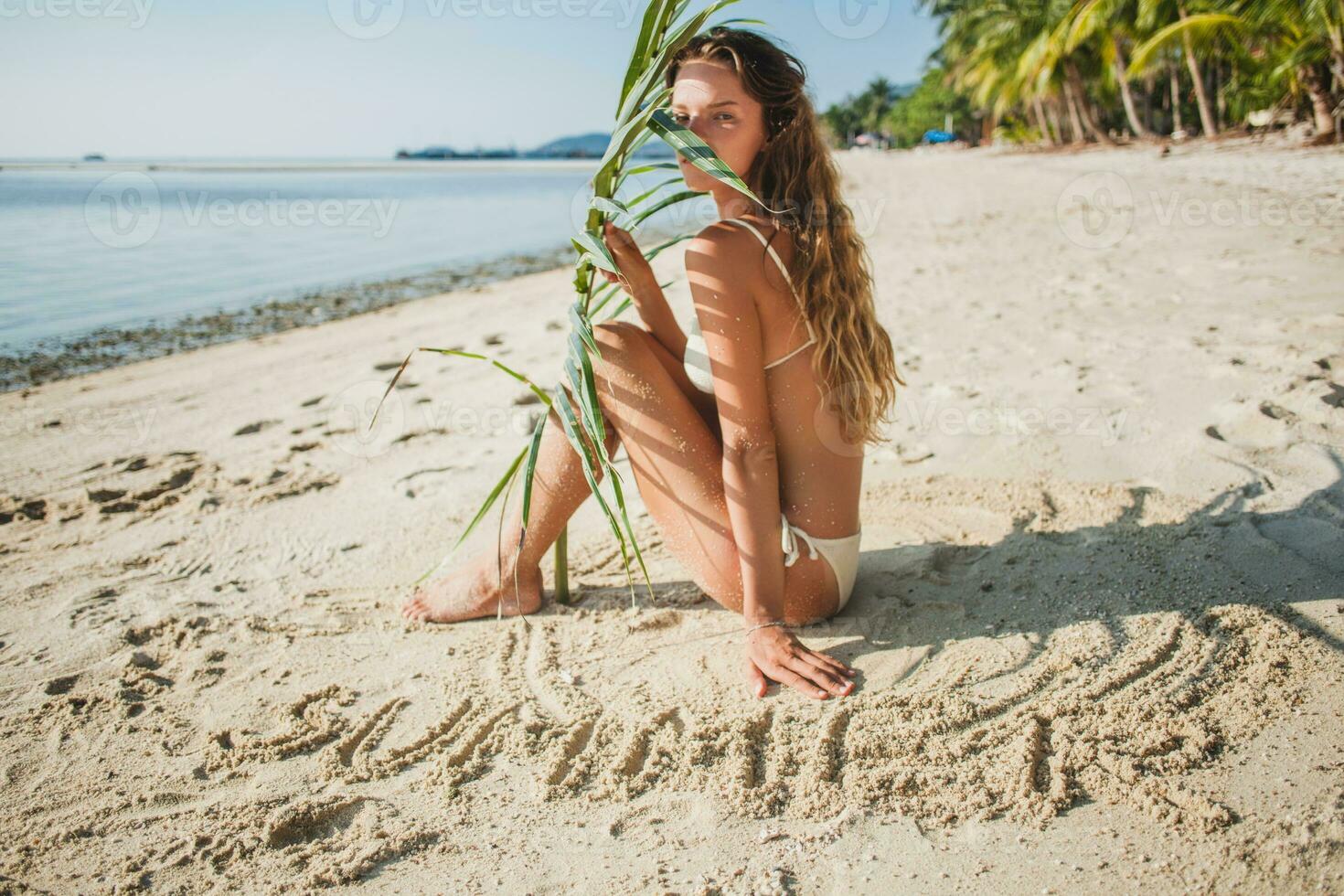 young skinny woman in white bikini swimwear holding leaf of palm tree photo