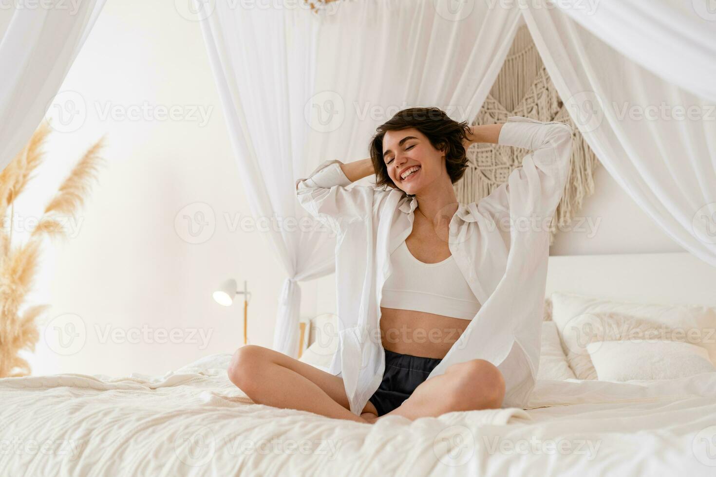 woman wake up in morning in boho style bedroom, sitting on bed lazy stretching, happy smiling photo