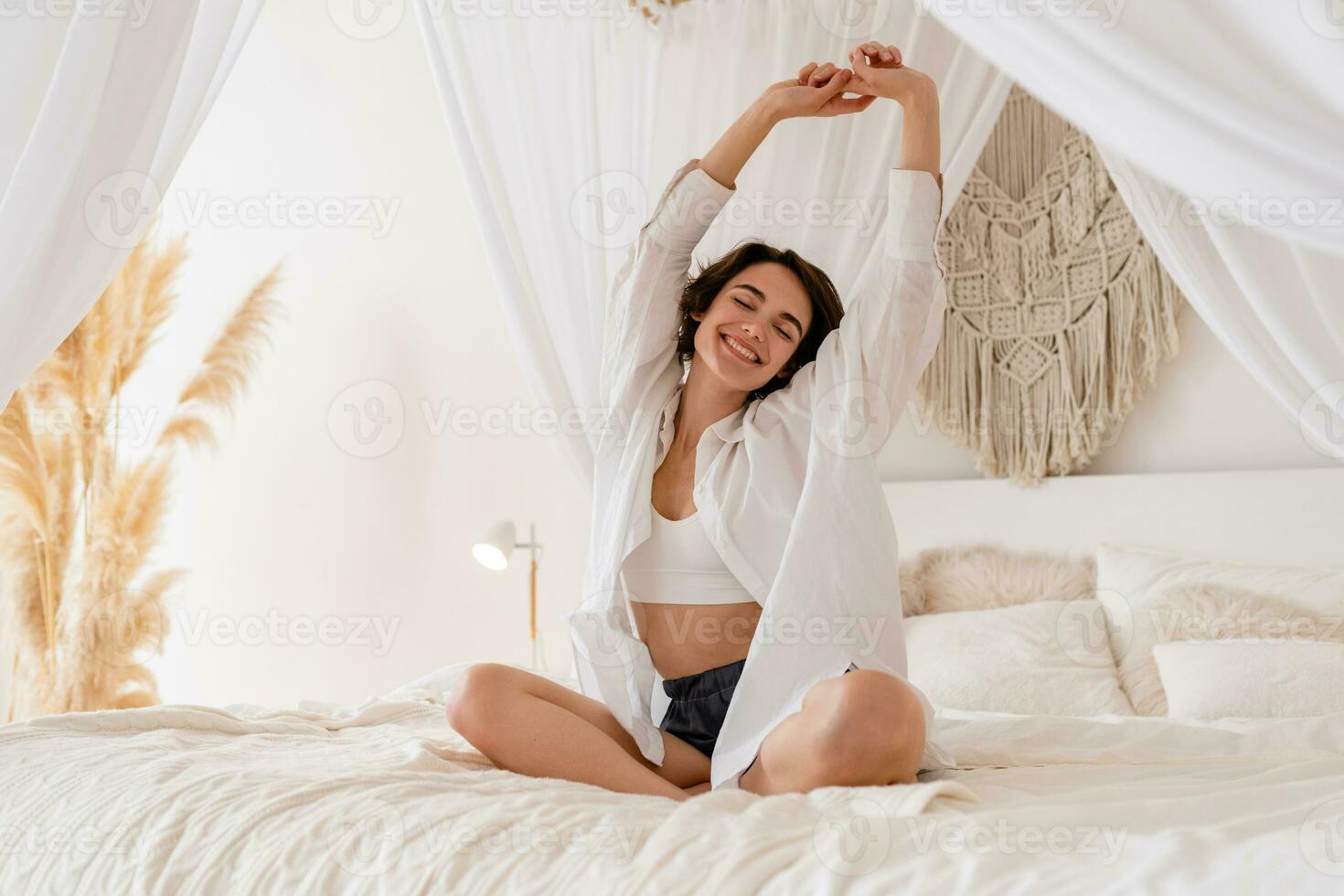 woman wake up in morning in boho style bedroom, sitting on bed lazy stretching, happy smiling photo