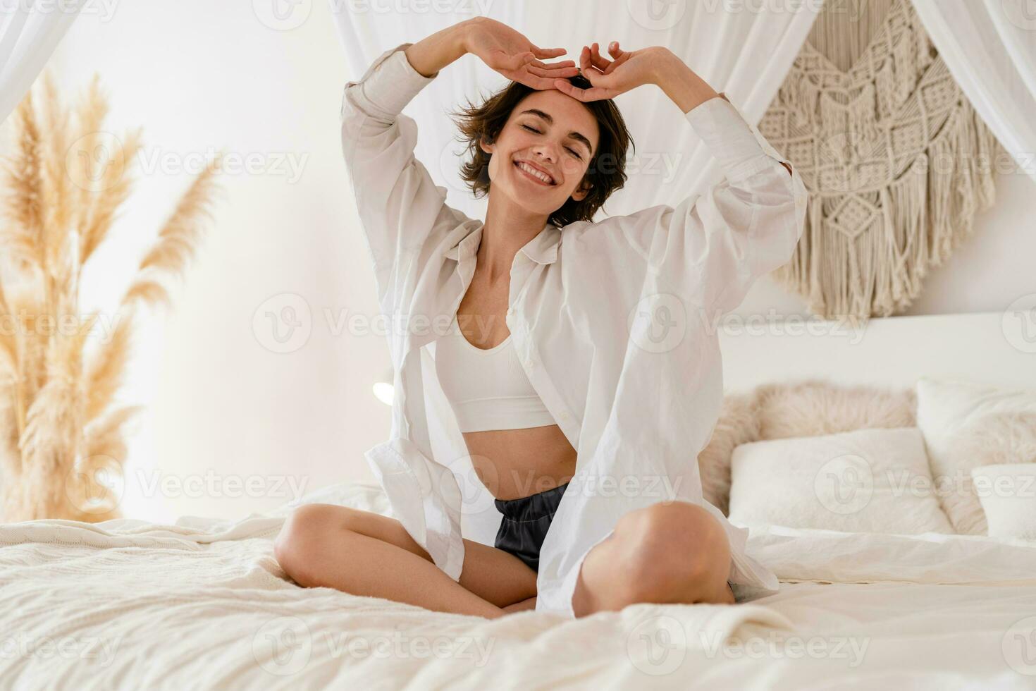 woman wake up in morning in boho style bedroom, sitting on bed lazy stretching, happy smiling photo