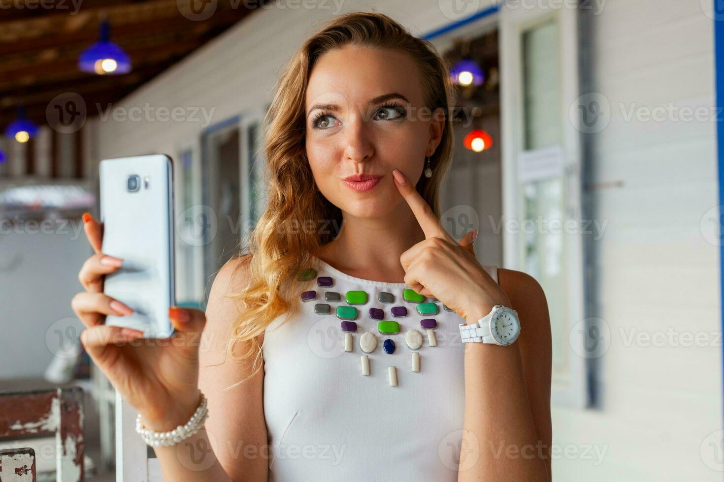 attractive woman in white dress in summer cafe in sunglasses using phone taking photo