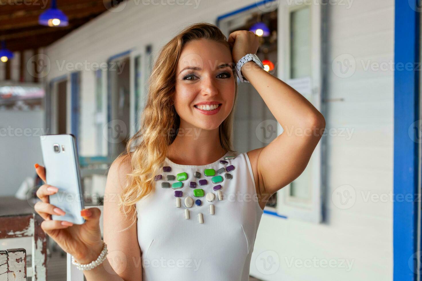 attractive woman in white dress in summer cafe in sunglasses using phone taking photo